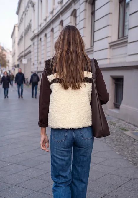 Eine Frau mit braunen Haaren, brauner Jacke und weißer Felltasche läuft eine gepflasterte Straße entlang.