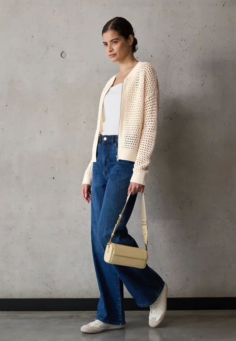 Vrouw met crèmekleurig vest, wit top, blauwe jeans en gele tas voor een betonnen muur.