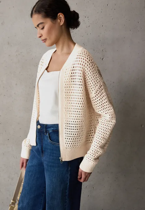 Vrouw met crèmekleurig vest, wit top en spijkerbroek voor betonnen muur.