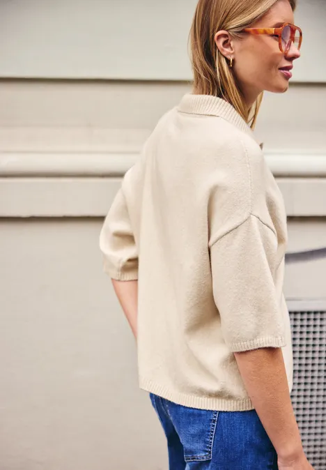 Kurzarm Cardigan mit Polokragen creamed sand Eine Frau trägt eine beige Bluse mit halblangen Ärmeln, blaue Jeans und orange Sonnenbrille, seitlich stehend.