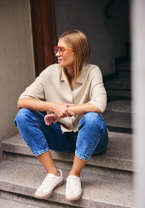 Kurzarm Cardigan mit Polokragen creamed sand Junge Frau in Jeans und Pullover sitzt lässig auf Treppenstufen und schaut zur Seite.