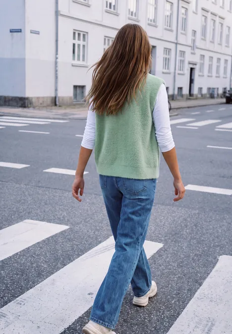 Eine Frau mit grünem Pullunder, weißen Shirt und Jeans überquert bei Tageslicht einen Zebrastreifen in der Stadt.