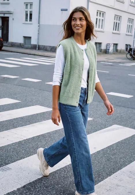 Frau in Jeans, weißem Shirt und grünem Strickwestchen überquert lächelnd bei sonnigem Wetter einen Zebrastreifen.