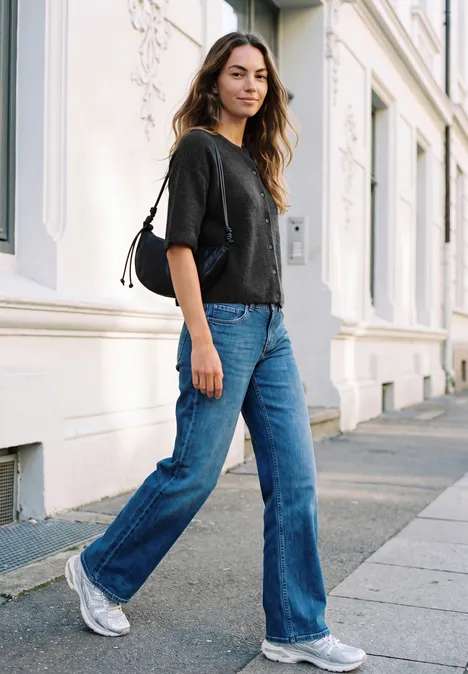 Frau in Jeans, schwarzem T-Shirt und weißen Sneakers geht entspannt eine sonnige Straße entlang.