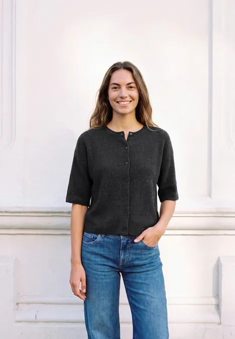 Eine junge Frau steht vor einer weißen Wand, trägt einen schwarzen Pullover und blaue Jeans und lächelt.