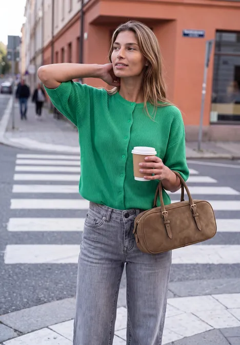 Frau in grünem Pullover mit Kaffee und Tasche wartet am Zebrastreifen.