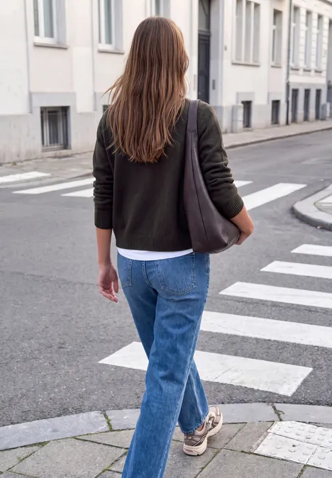 Eine Frau geht auf einem Zebrastreifen in der Stadt, trägt Jeans, Pullover und eine Tote Bag.