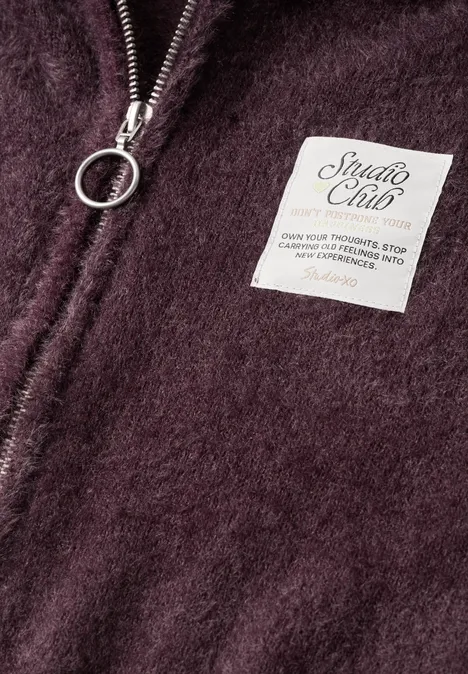 Weste mit Kapuze und Zipper very berry Nahaufnahme eines weinroten Fleece-Teils mit Reißverschlussring und weißem 'Studio Club'-Label.