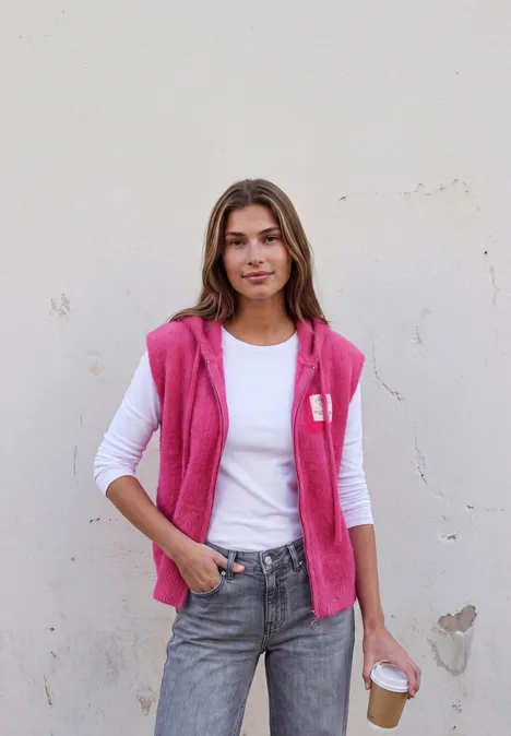 Junge Frau mit weißem T-Shirt und pinker Weste, lässig in Jeans, hält einen Kaffee vor einer hellen Wand.