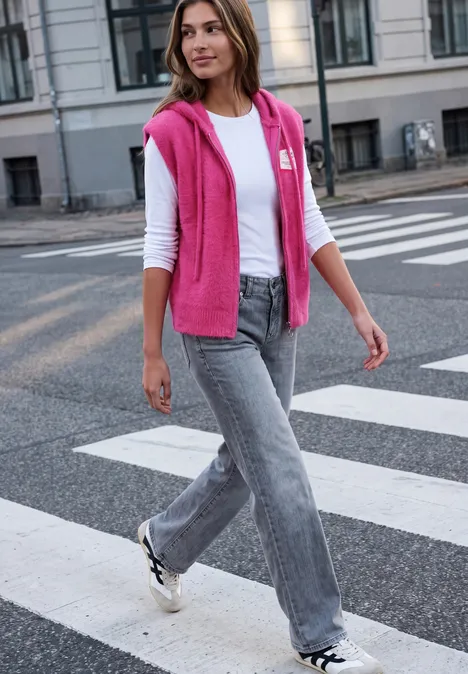Frau in weißem Shirt, grauen Jeans und pinkem Pulli über den Schultern läuft über Zebrastreifen.