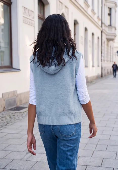 Frau in grauer Weste und Jeans geht auf einer gepflasterten Straße, Blick von hinten.