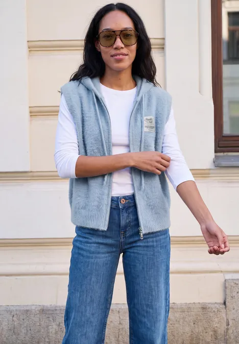 Eine Frau in Jeans, weißem Langarmshirt und ärmelloser Kapuzenweste steht vor einem Gebäude mit Sonnenbrille.