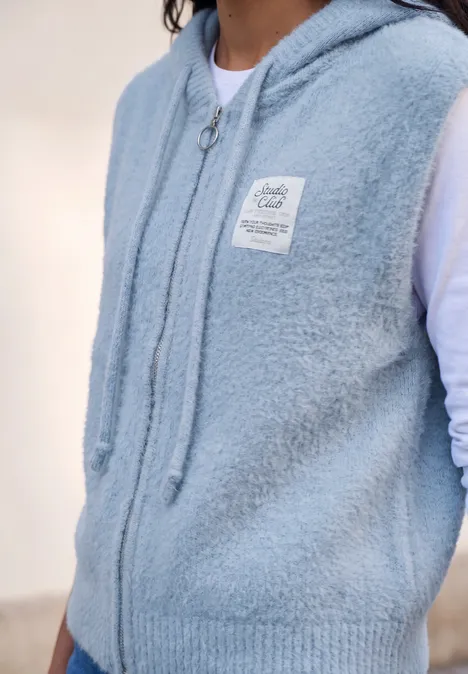 Hellblaue, flauschige Kapuzenweste mit Reißverschluss und Brustlabel, darüber ein weißes Langarmshirt.