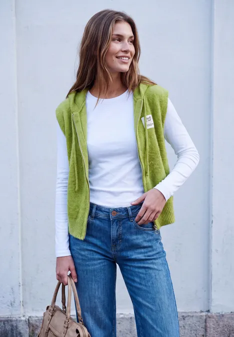 Vrouw met witte blouse, groen vest, jeans en tas, lachend voor een lichte achtergrond.