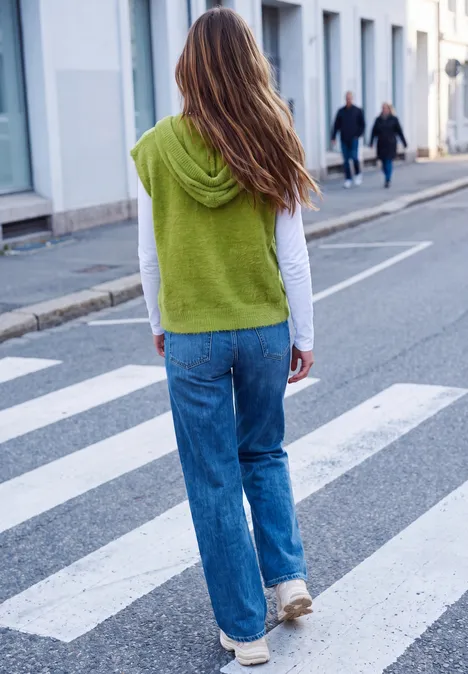 Vrouw in jeans en wit topje, groene trui over de schouders, loopt over een zebrapad.