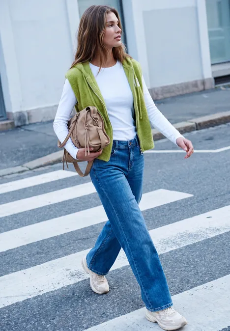 Een vrouw met wit shirt, groen vest, wijdere jeans en beige tas steekt een zebrapad over in de stad.