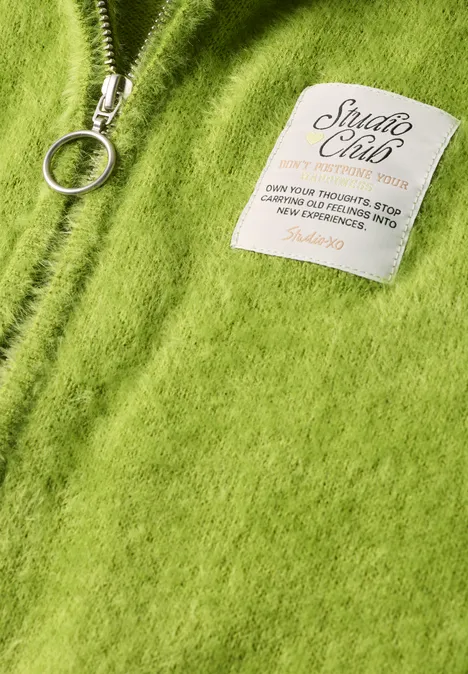 Close-up van een zacht, grasgroen wollen vest met zilverkleurige rits en 'Studio Club'-label.