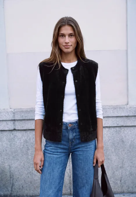 Eine junge Frau in schwarzer Weste, weißem Shirt und Jeans trägt eine schwarze Tasche und lächelt.