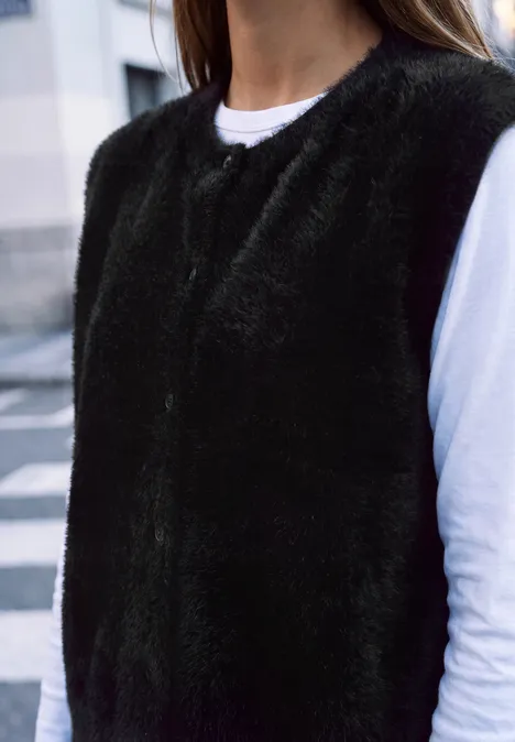 Weibliche Person trägt schwarze, flauschige Weste über weißem Langarmshirt.