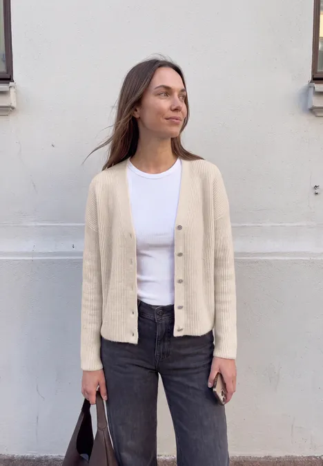 Cardigan mit V-Neck und Knopfleiste creamed sand mel. Junge Frau lehnt an weißer Wand, trägt beigen Cardigan, weißes Shirt und schwarze Jeans, hält schwarze Tasche.