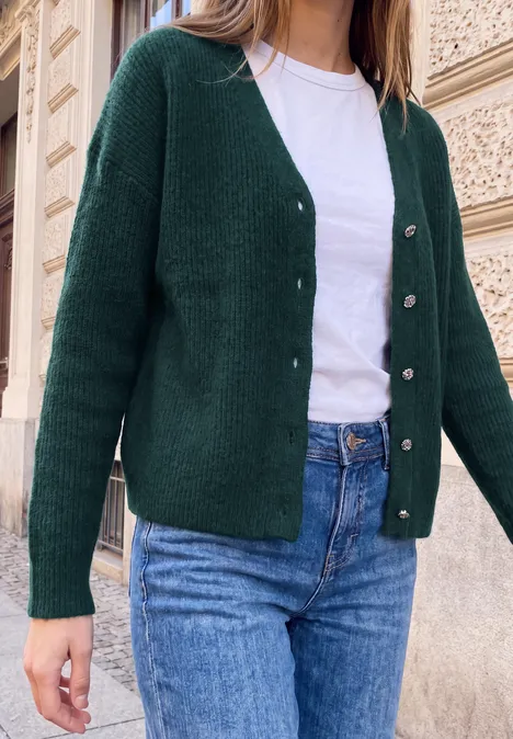 Cardigan mit V-Neck und Knopfleiste mossed green mel. Frau trägt grünen Cardigan, weißes Shirt und blaue Jeans vor einer steinernen Hausfassade.