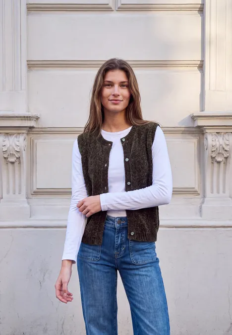 Strickweste mit Rundhals und Knöpfen charred brown Frau lehnt lächelnd an einer Fassade, trägt weißes Shirt, Weste und Jeans.