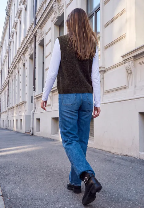 Strickweste mit Rundhals und Knöpfen charred brown Eine Person mit blauer Jeans, schwarzer Weste und weißem Longsleeve spaziert an einem weißen Gebäude vorbei.