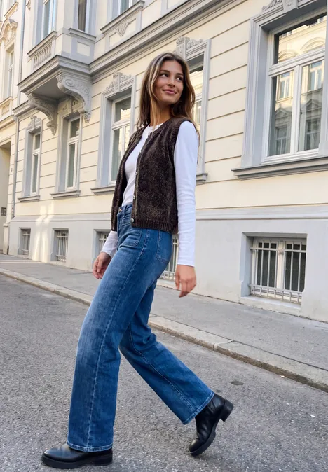 Strickweste mit Rundhals und Knöpfen charred brown Junge Frau in Jeans, weißem Shirt und brauner Weste geht lächelnd an elegantem Stadthaus vorbei.