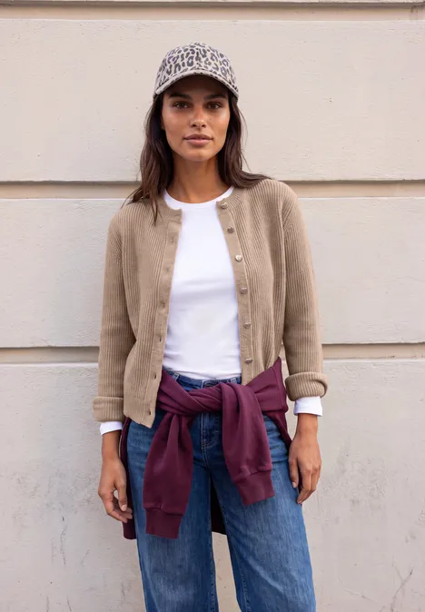 Cardigan à côtes et col rond biscuit sand mel. Eine junge Frau vor heller Wand, trägt beige Jacke, weißes Shirt, Jeans, lila Pullover um die Taille und ein gemustertes Cap.