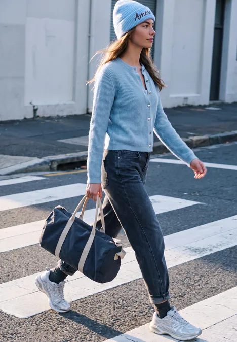 Cardigan à côtes et col rond loom blue mel. Eine Frau überquert einen Zebrastreifen, trägt blaue Mütze, Jacke, dunkle Hose und weiße Sneaker, hält Tasche.