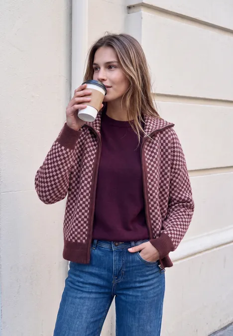 Frau in Jeans und karierter Jacke trinkt Kaffee aus Pappbecher und lehnt an heller Wand.
