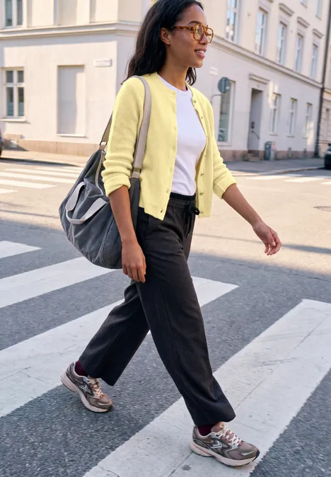 Cardigan à côtes et col rond healing yellow mel. Frau mit gelber Jacke und Rucksack überquert bei Sonnenschein einen Zebrastreifen in der Stadt.