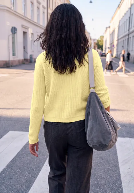Cardigan à côtes et col rond healing yellow mel. Frau mit gelbem Pullover und grauer Tasche überquert bei Sonnenschein einen Zebrastreifen.