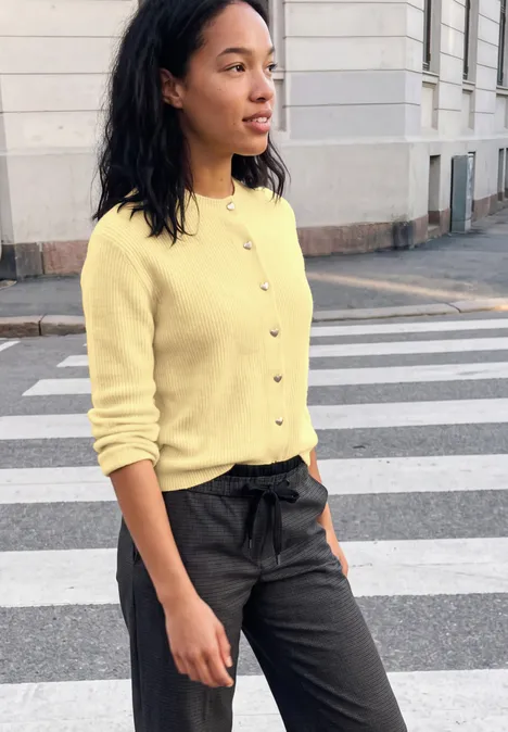 Cardigan à côtes et col rond healing yellow mel. Eine Frau in gelbem Cardigan und schwarzen Hosen überquert einen Zebrastreifen in der Stadt.