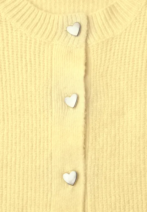 Cardigan à côtes et col rond healing yellow mel. Nahaufnahme eines gelben Strickoberteils mit silbernen herzförmigen Knöpfen.
