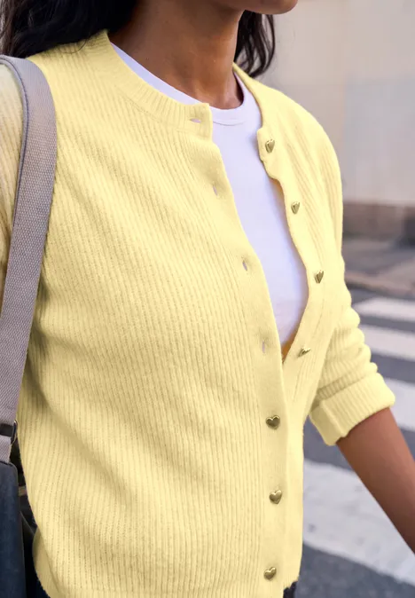 Cardigan à côtes et col rond healing yellow mel. Eine Frau trägt eine gelbe Strickjacke über einem weißen Shirt.