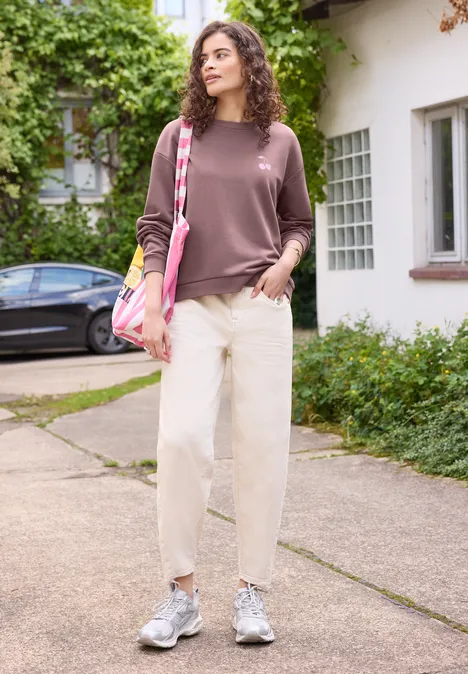 Une femme en tenue décontractée avec pantalon clair, baskets grises et sac rose devant une maison.