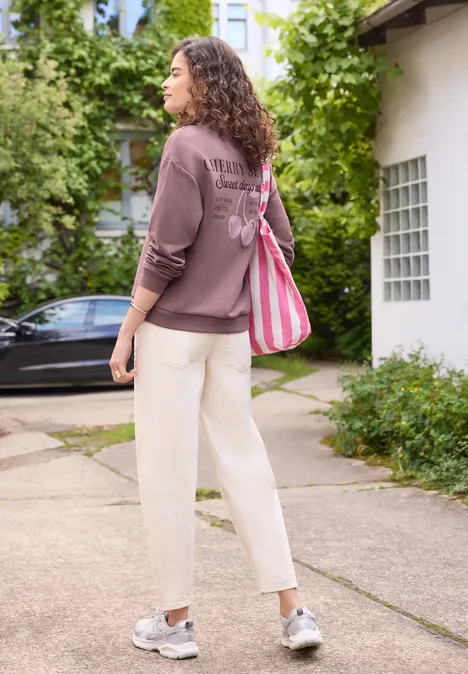 Femme en pull marron, pantalon blanc et sac rayé marche sur un trottoir bordé de végétation.