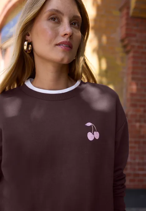 Personne en sweat marron avec motif cerises roses devant un mur.