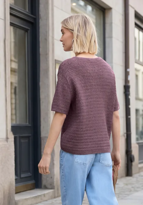 Eine Frau mit kurzen blonden Haaren in violettem Strickshirt und blauer Jeans geht eine gepflasterte Straße entlang.