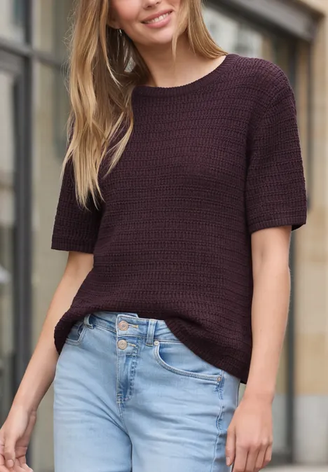Frau in dunkelviolettem Kurzarm-Pullover und hellblauer Jeans, stehend vor einer Hausfassade.