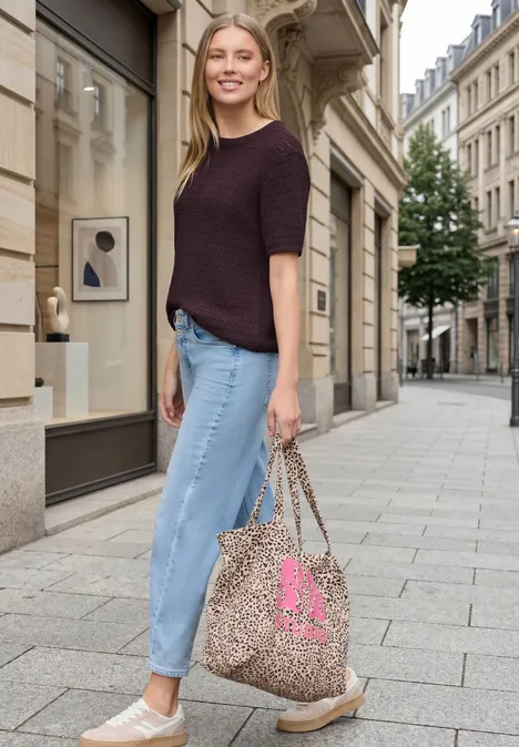 Frau in lässigem Outfit mit Strickshirt, hochgekrempelten Jeans, Slip-ons und Leoparden-Print-Tasche in Stadtgasse