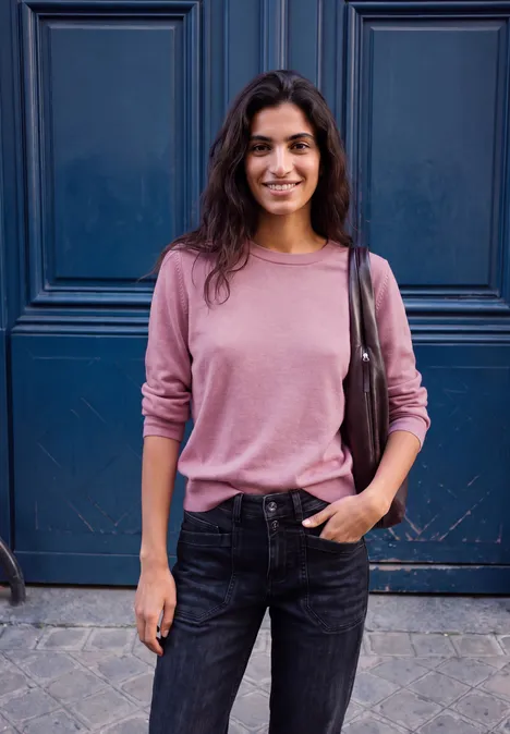 Eine lächelnde Frau in rosa Pullover und schwarzen Jeans steht vor einer blauen Tür.