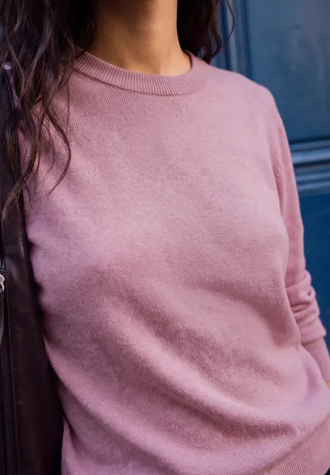Frau mit langen dunklen Haaren trägt rosa Pullover vor dunkler Tür.