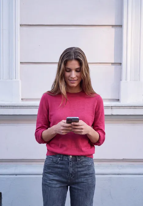 Pullover mit Rundhals in Unifarbe ruby raspberry mel. Eine junge Frau steht vor einer Hauswand und schaut auf ihr Smartphone.