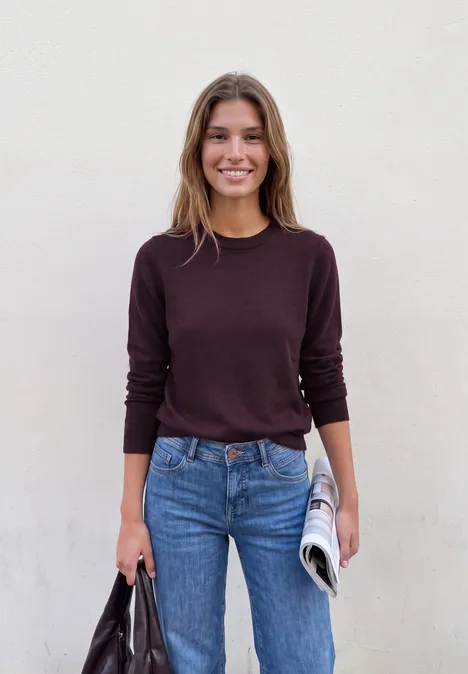 Lächelnde Frau in dunklem Pullover und Jeans steht vor weißer Wand, hält Zeitung und Tasche.