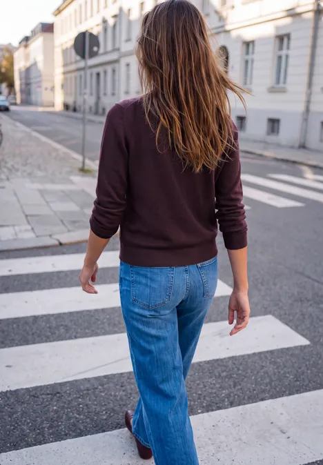 Frau im braunen Pulli und Jeans überquert zu Fuß einen Zebrastreifen in einer städtischen Straße.