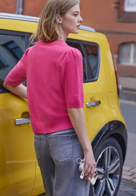 Frau mit pinkem Oberteil und Jeans hält eine Sonnenbrille neben einem gelben Auto in urbaner Umgebung.