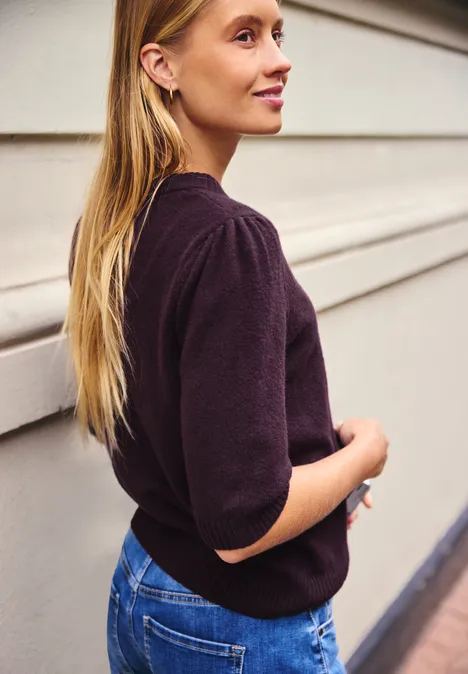 Jeune femme aux cheveux blonds longs, appuyée de profil contre un mur, portant un pull sombre et un jean.