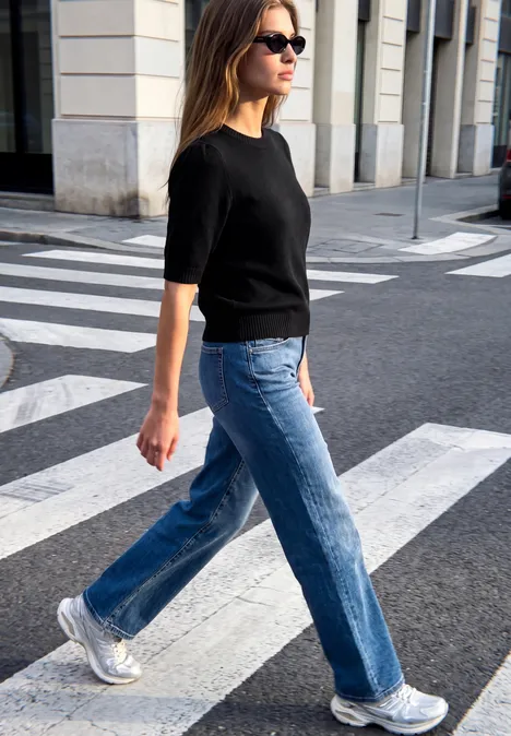 Frau mit Sonnenbrille, schwarzem Pullover, blauen Jeans und silbernen Sneakern überquert Straße.
